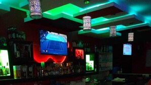 A bar area with red and black bar stools, a large screen displaying a game, and green ambient lighting. - Nestwood Hotel (Nanyuki)