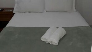 Cama bem arrumada com manta cinza e duas toalhas enroladas, cabeceira branca e mesa de cabeceira de madeira com controle remoto e um livro.