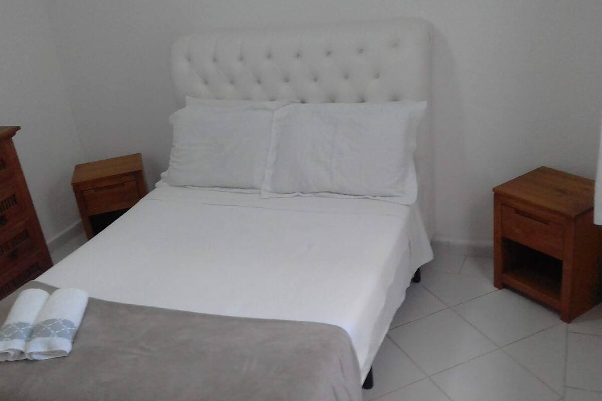 Un lit bien fait, avec du linge de lit blanc, dans une pièce simple aux murs et aux meubles en bois, le tout d’une grande sobriété.