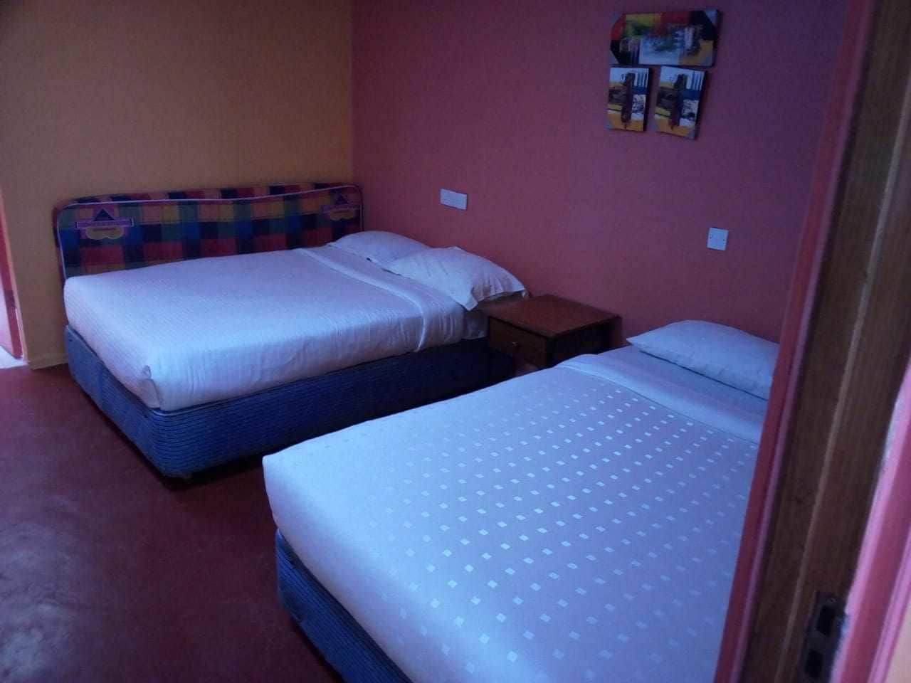 Ein Zimmer mit zwei Betten, jedes mit einem anderen Kopfteil. Dazwischen steht ein kleiner Tisch, darüber eine Wandregal.