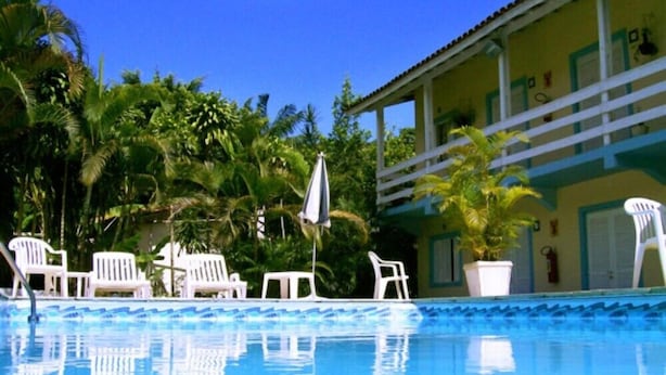 A swimming pool with white lounge chairs and umbrellas, surrounded by lush greenery and a building with a balcony. - Apartamentos Atobá Juquehy (São Sebastião)