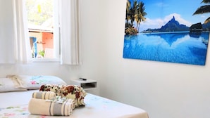 A bedroom with a bed, a window with curtains, a painting of a tropical scene, and a small bedside table. - Apartamentos Atobá Juquehy (São Sebastião)