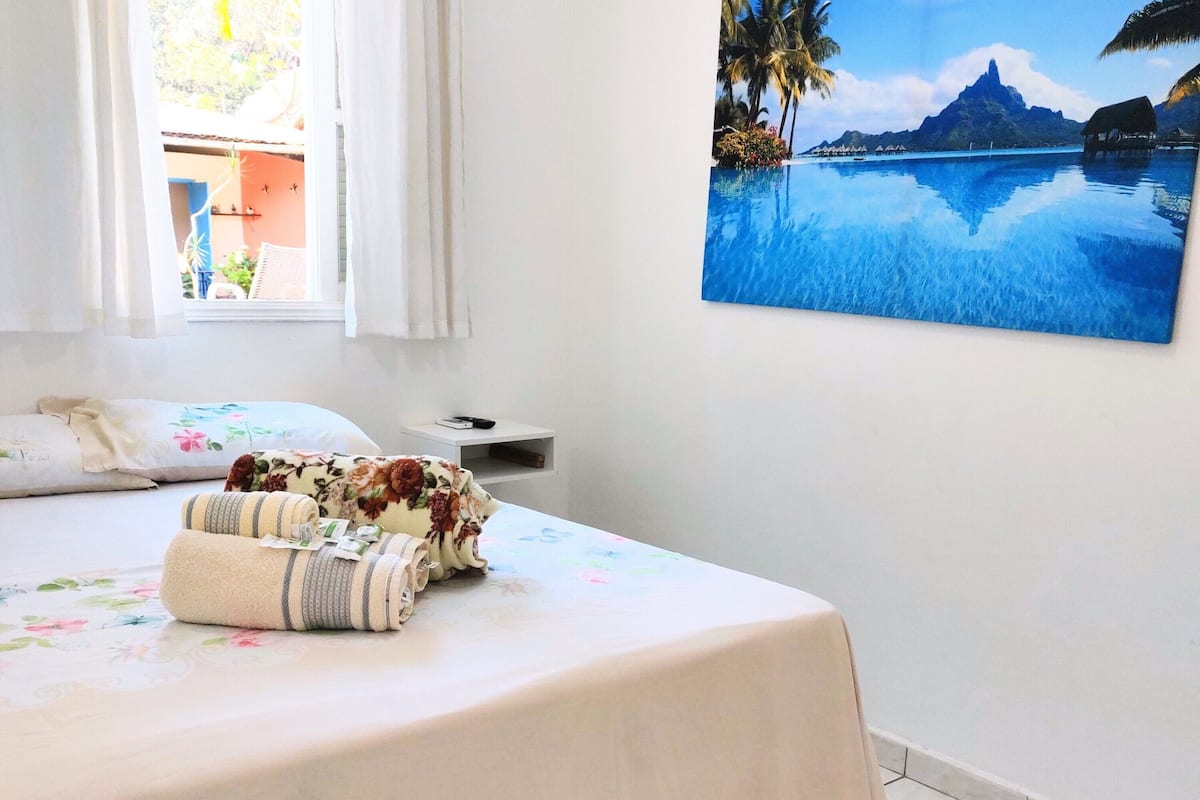 Une chambre à coucher avec un lit, une fenêtre avec des rideaux, un tableau représentant un paysage tropical et une petite table de chevet.