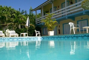 Piscina con sillas de descanso blancas y sombrillas, rodeada de palmeras y un edificio con detalles azules y blancos.