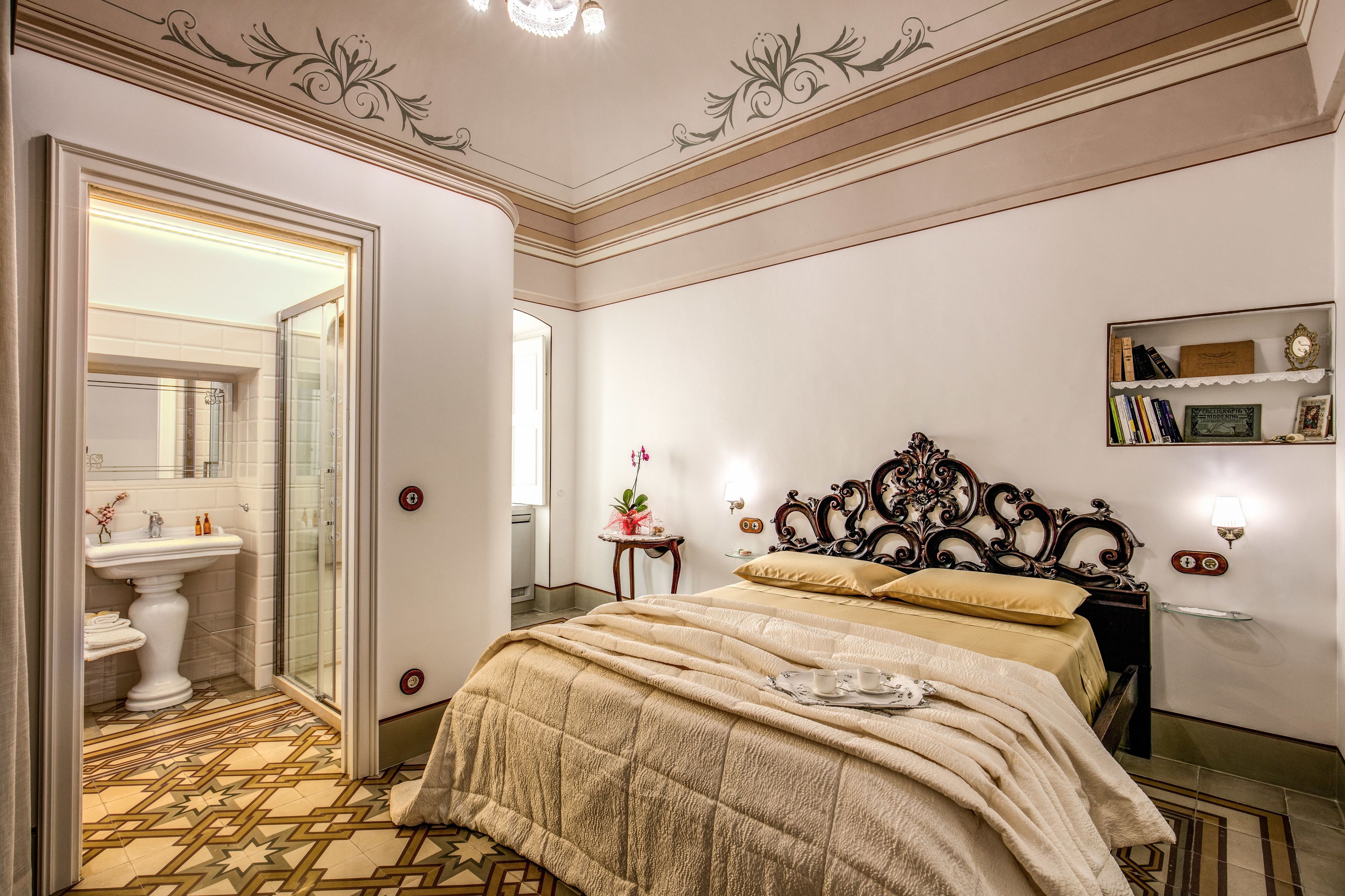 Une chambre d’hôtel avec un grand lit, une table de chevet avec une lampe, une petite table d’appoint avec une plante, et une salle de bain avec un lavabo et un miroir.