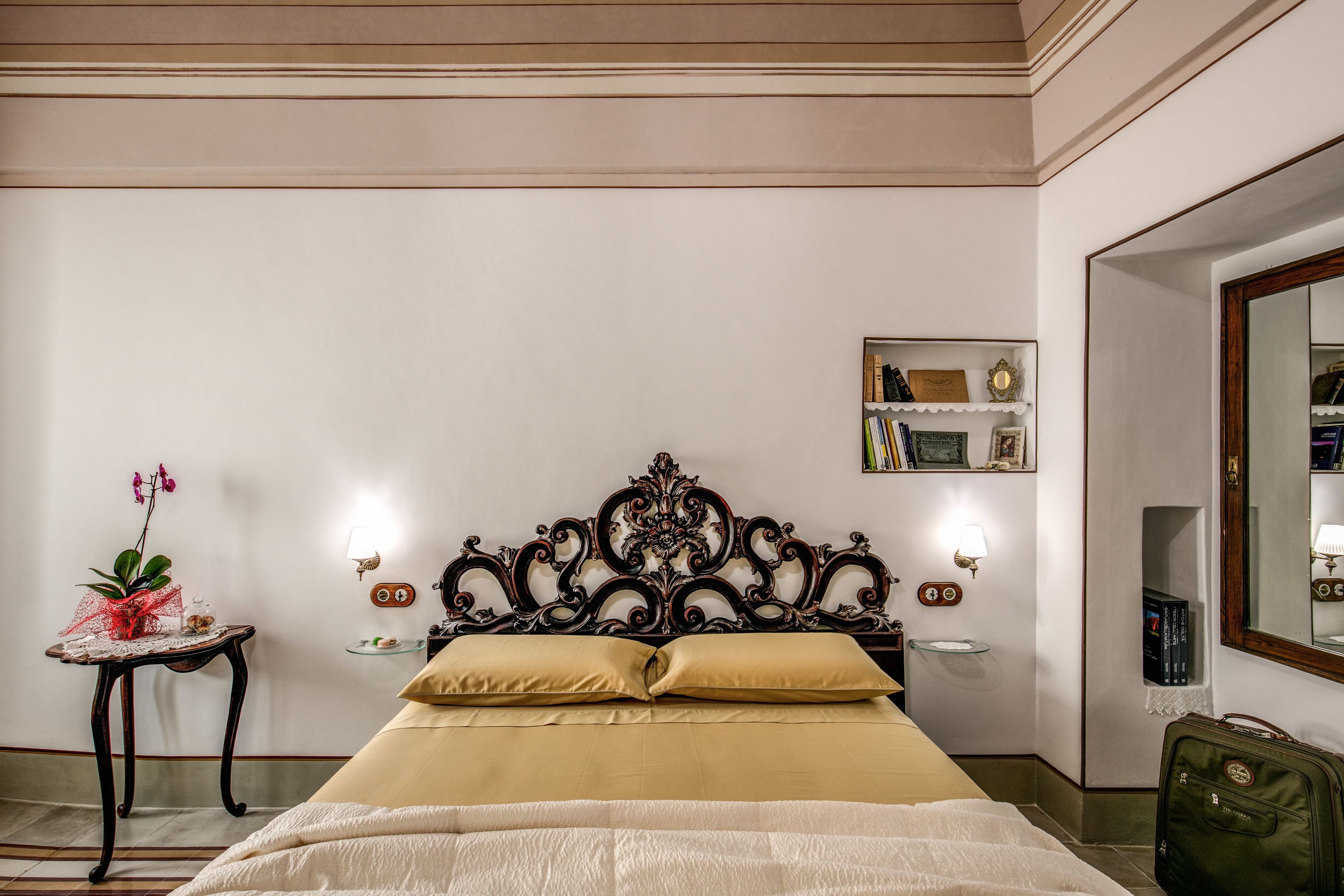 Quarto de hotel com uma cama grande, uma mesa de cabeceira com abajur, uma pequena estante com livros e um espelho na parede.