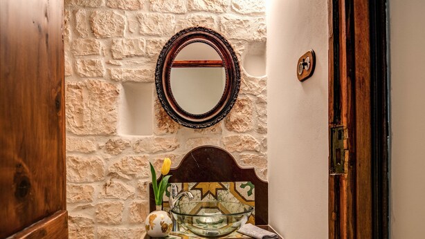 A bathroom with a stone wall, a wooden door, a round mirror, a glass bowl sink, and a vase with flowers. - Palazzo Stunis- Dimora di Charme (Ostuni)