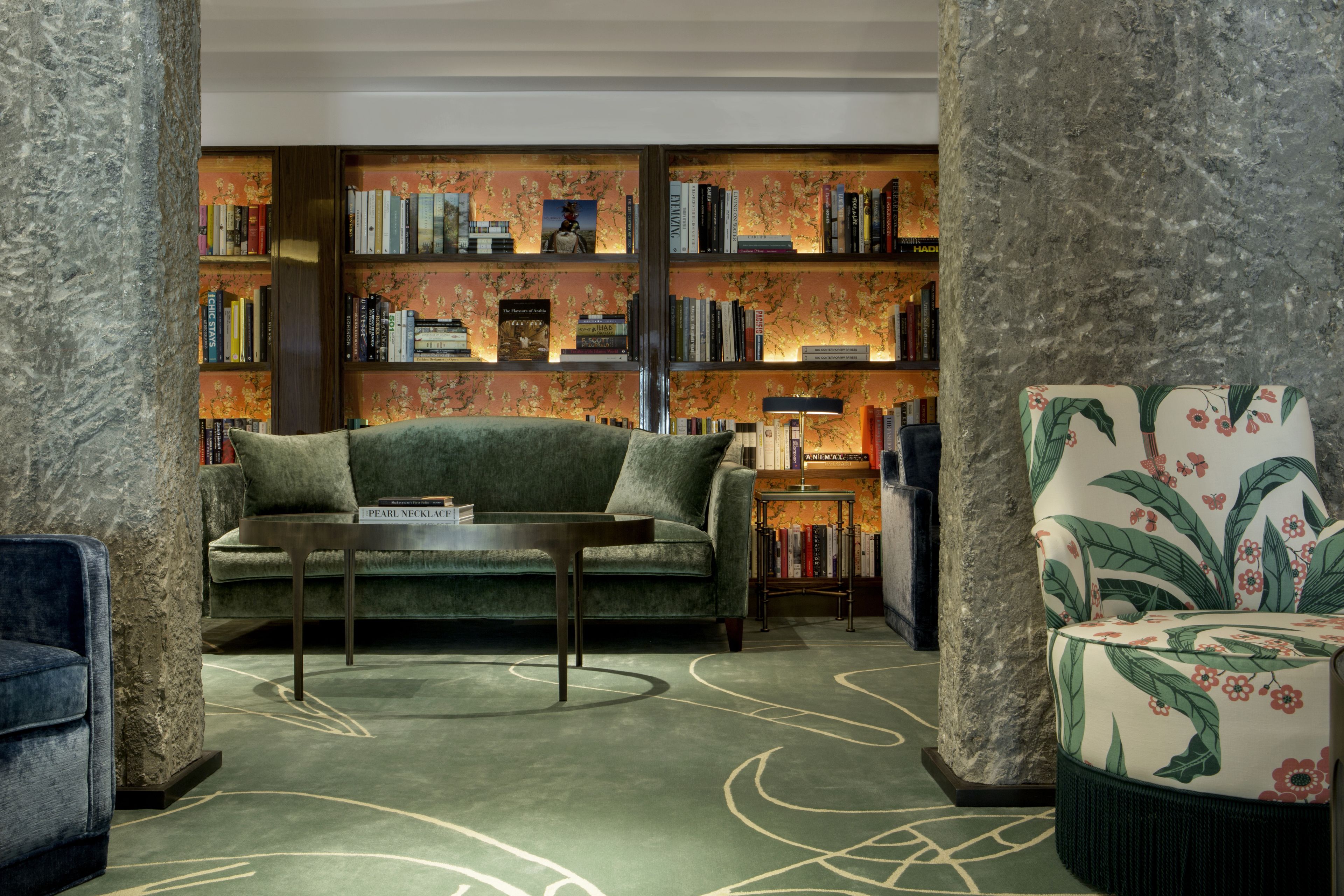 Un salón acogedor con un sofá verde, un sillón estampado y una estantería llena de libros.
