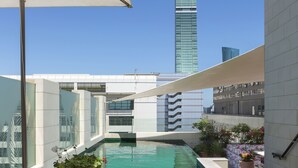 Una piscina en la azotea con vistas a un rascacielos alto, rodeada de edificios modernos y equipada con sillones y una mesa.