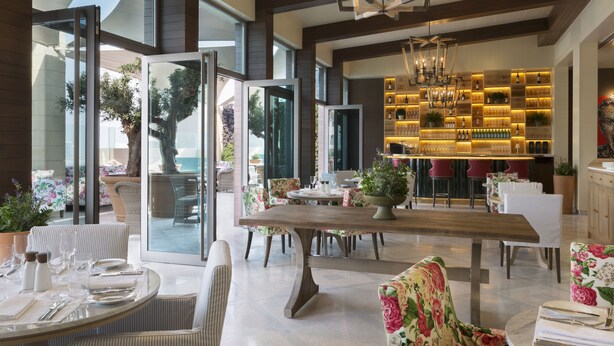 Un comedor amplio con una gran mesa de madera, rodeado de sillas con motivos florales, y una zona de bar con estantes llenos de botellas y vasos.
