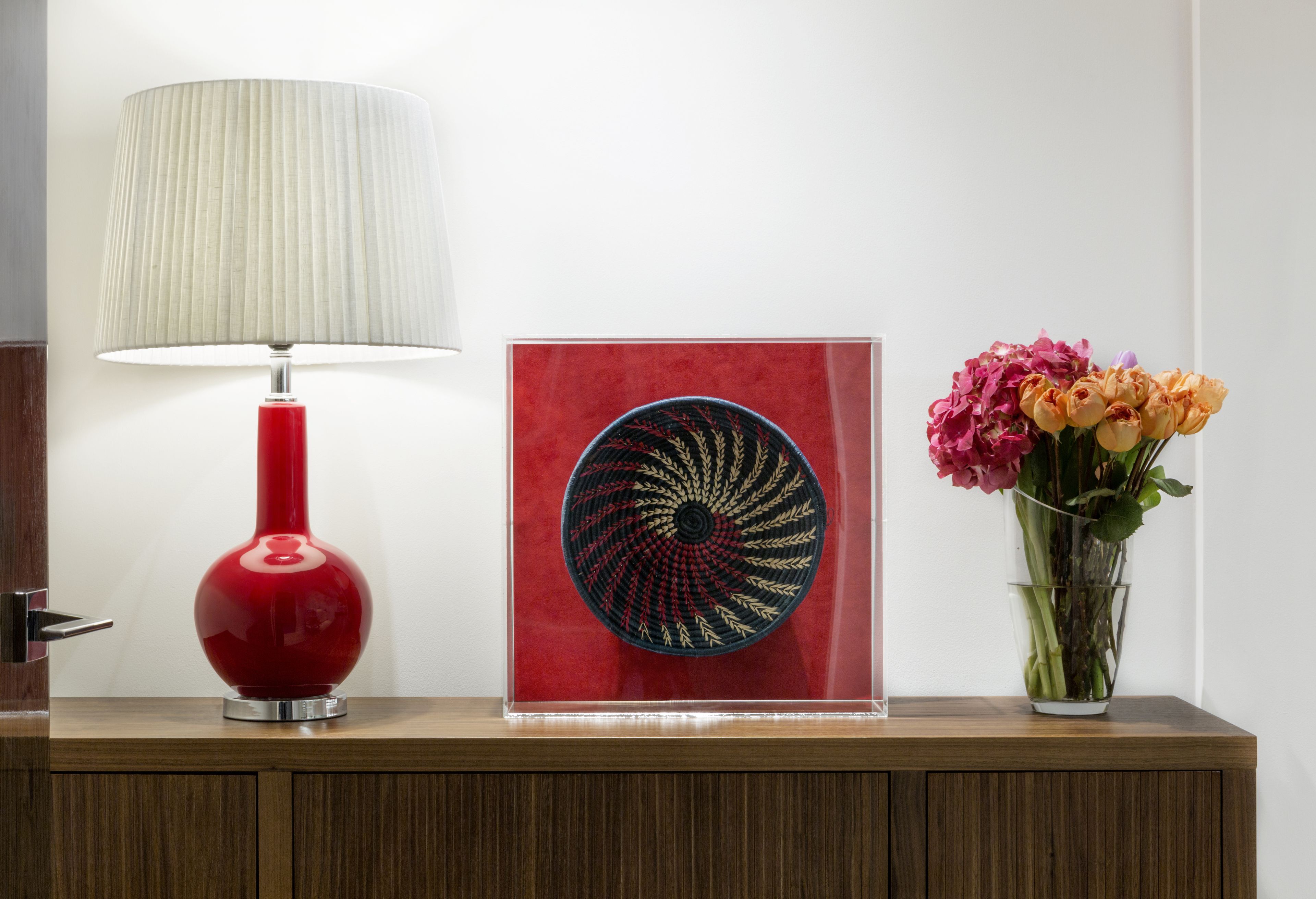 A red lamp, a framed artwork, and a vase with flowers on a wooden surface.