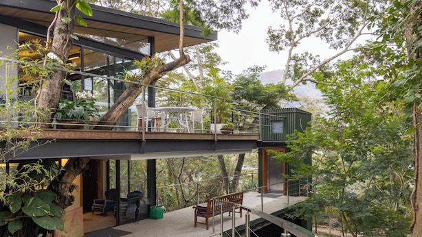 A modern house with a balcony, outdoor seating, and lush greenery.