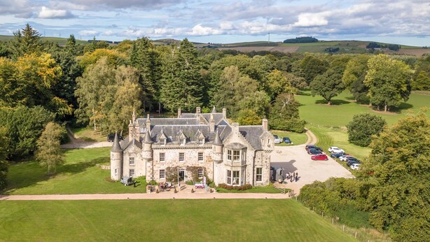 A large stone building with multiple towers, surrounded by lush greenery and a parking area with several cars. - Wardhill Castle (Inverurie)