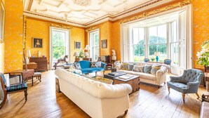 A spacious living room with a white sofa, blue armchair, and a coffee table. There are large windows providing natural light and a view of greenery outside. - Wardhill Castle (Inverurie)