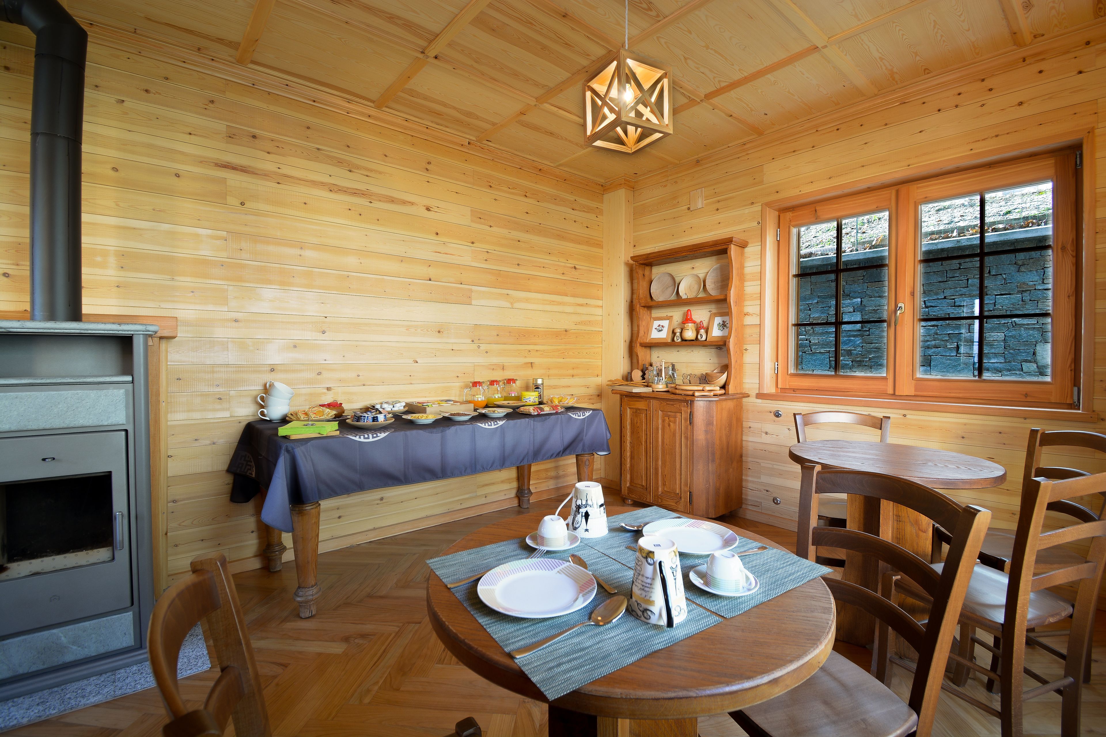 Interior de una cabaña de madera con una mesa de comedor preparada para el desayuno, una chimenea de piedra y una ventana con vistas.