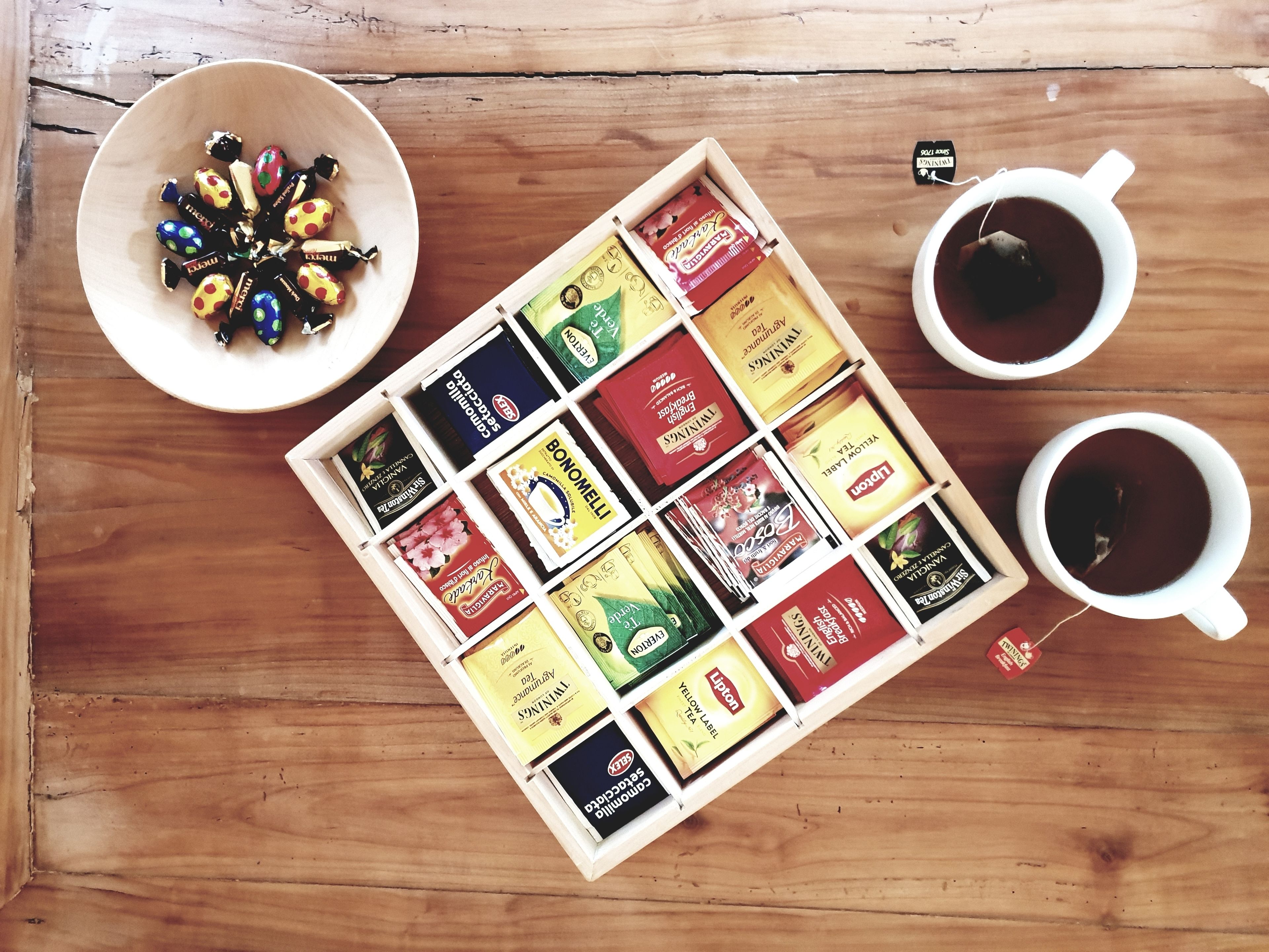 A wooden table with a bowl of colorful candies, two cups of tea, and a tray of assorted tea bags.