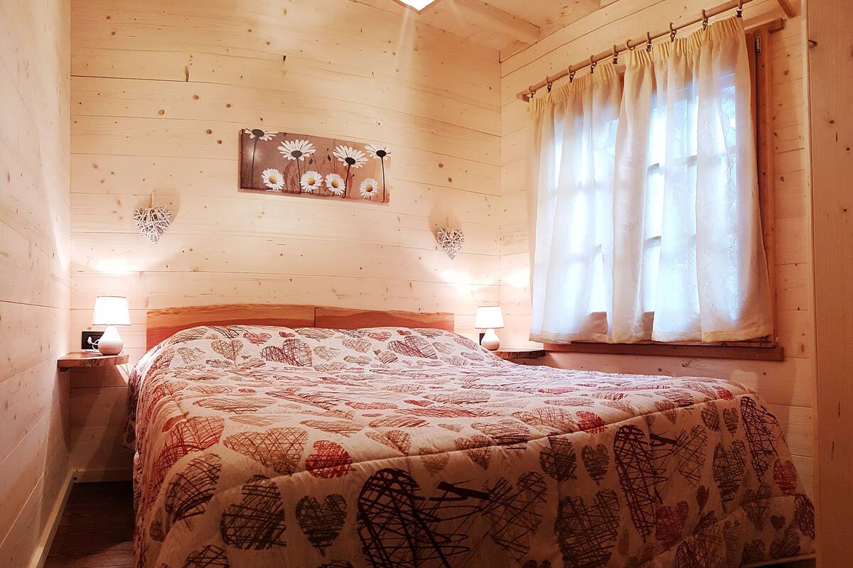 Ein gemütlicher Holzraum mit einem Bett, zwei Nachttischlampen, einem Fenster mit Vorhängen und einer Wanddekoration mit Gänseblümchen.