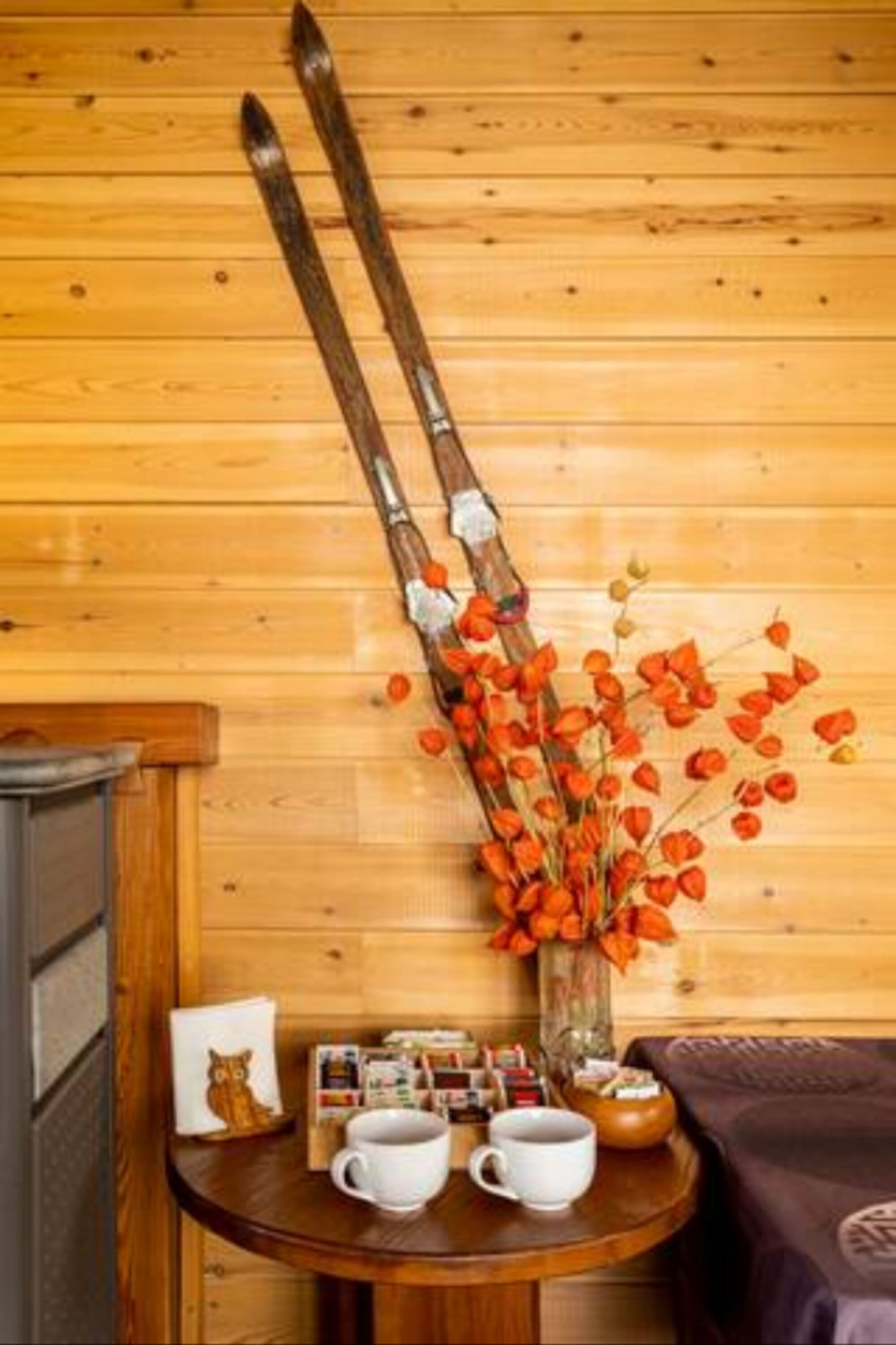 A wooden wall with a pair of skis leaning against it, a vase with orange flowers, and a table with two cups and a small bowl.