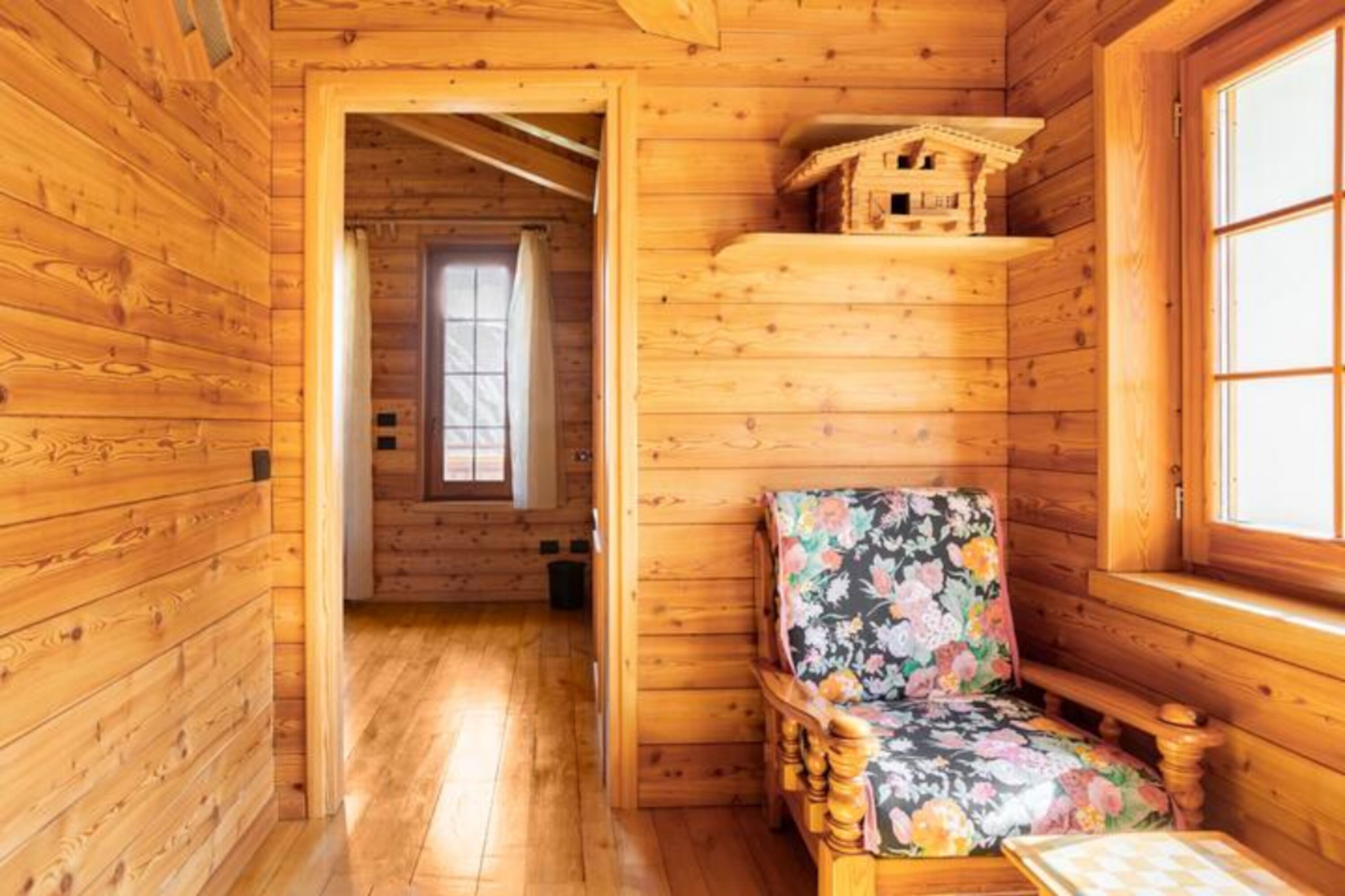 A wooden cabin interior with a wooden bench, a small house model on a shelf, and a window.