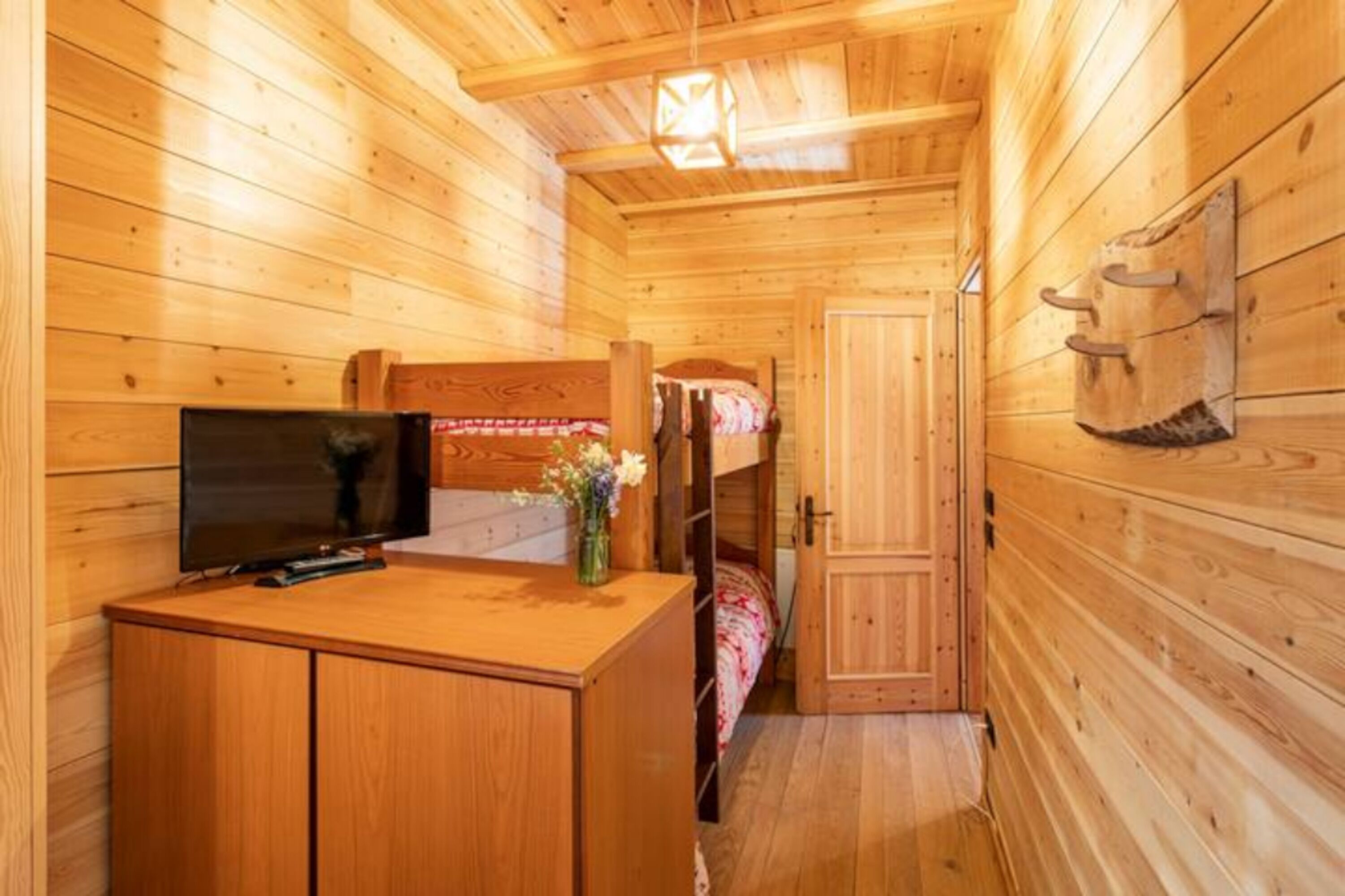 Interior de una cabaña de madera con una litera, un escritorio con televisor y una mesita con un florero.