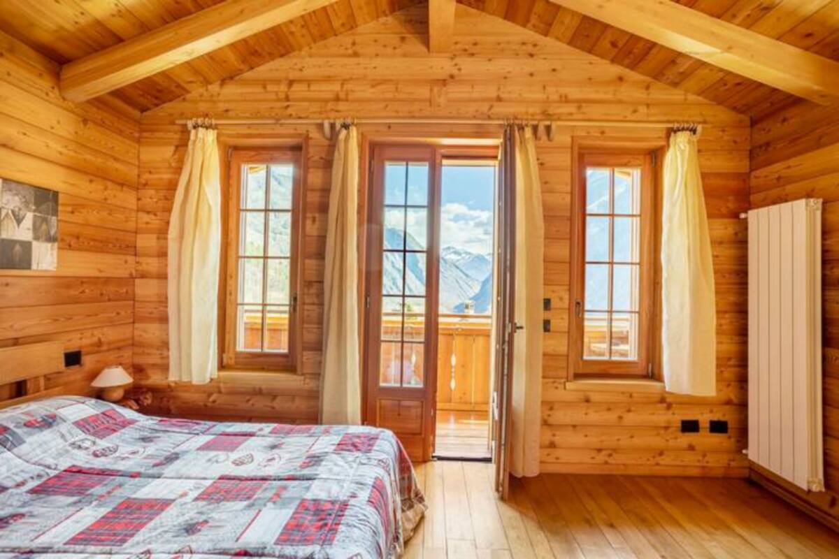 Ein Schlafzimmer einer Holzhütte mit Bett, zwei Fenstern, einer Tür und einem Heizkörper.