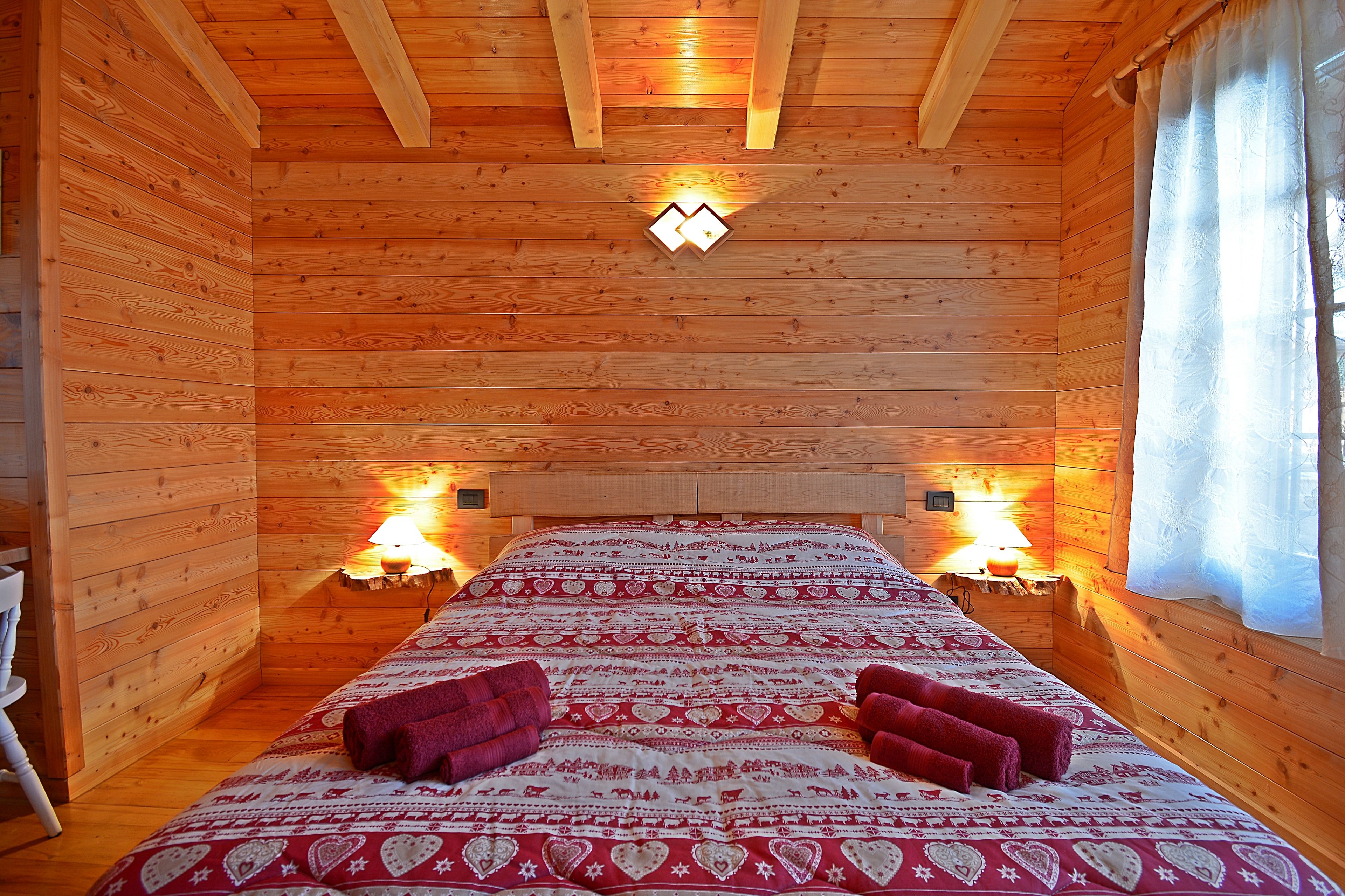 A wooden cabin room with a bed, two bedside lamps, and a window with curtains.
