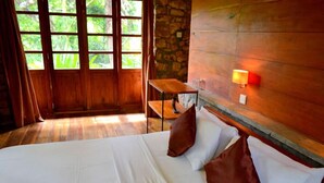 A hotel room with a wooden floor, a bed with white bedding and brown pillows, a small wooden table, a stone wall, and a window with curtains. - Landa Holiday House (Imbulpe)