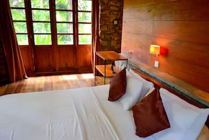 Habitación de hotel con piso de madera, cama con ropa blanca y almohadas marrones, una pequeña mesa de madera, pared de piedra y una ventana con cortinas.