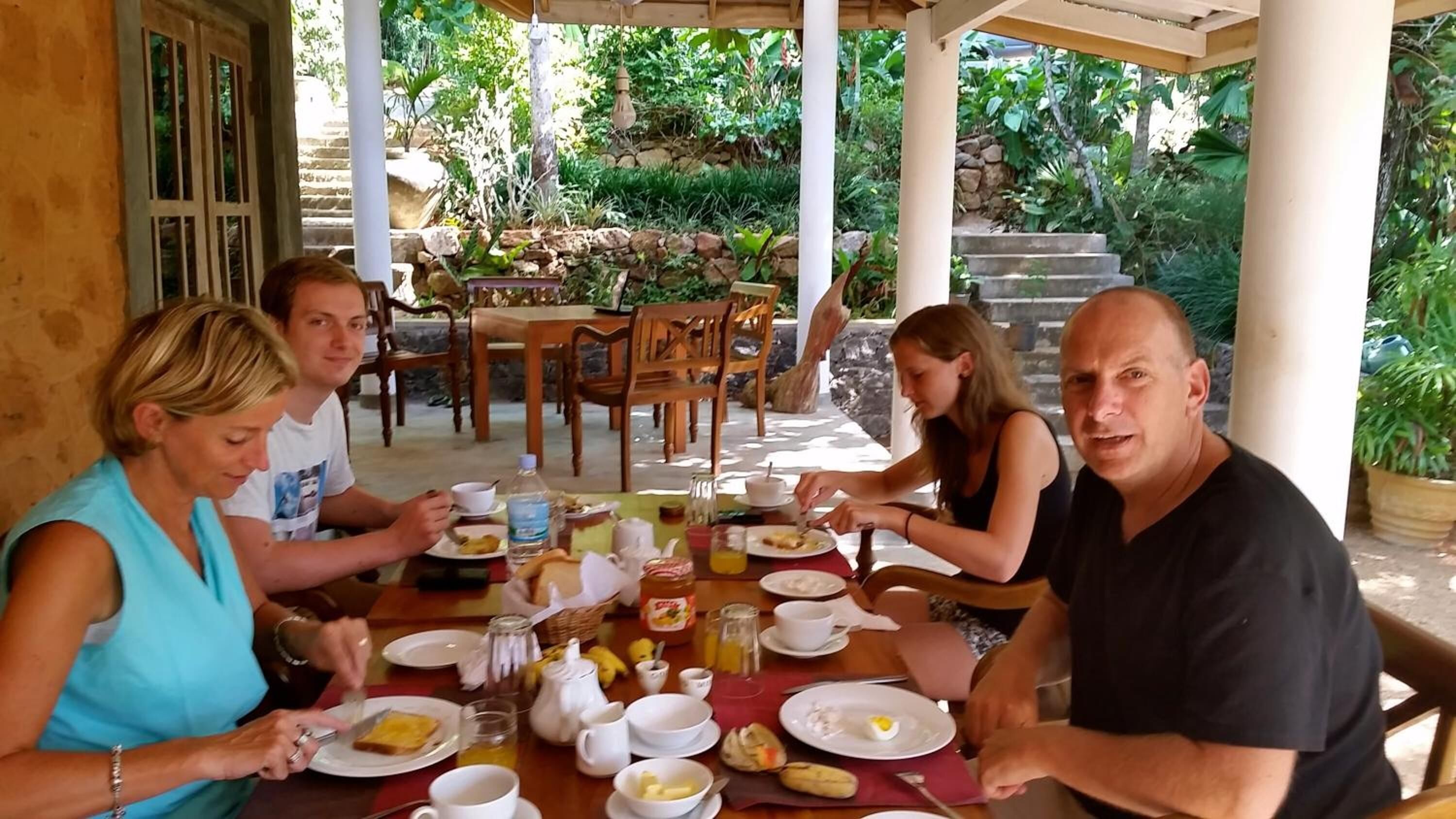 Un groupe de personnes est assis à une table en plein air et déguste divers plats, boissons servies dans des tasses, ainsi qu’une bouteille de jus de fruits.