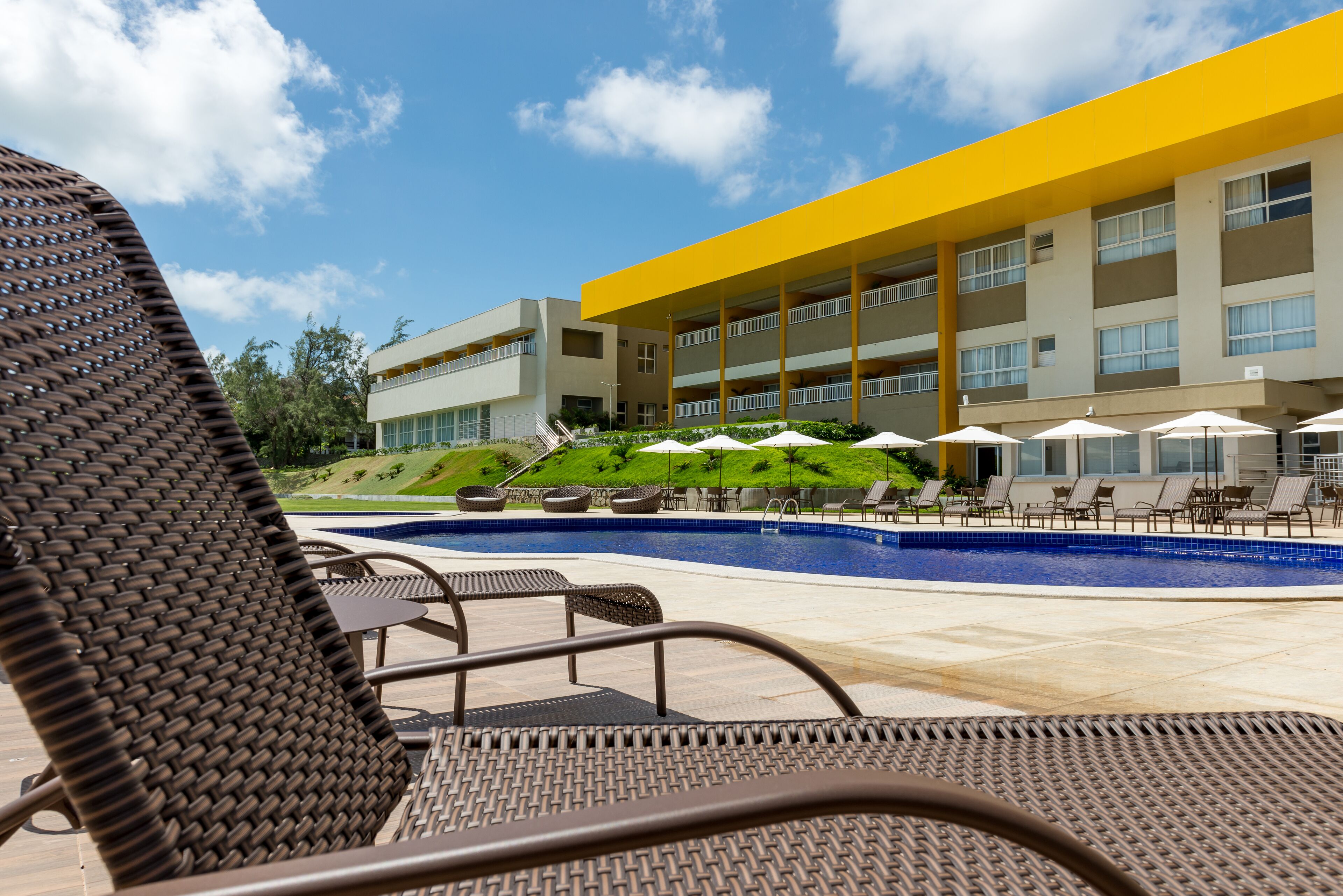 Ein Poolbereich mit Liegestühlen und Sonnenschirmen, ein modernes Gebäude mit gelben Akzenten und ein bepflanzter Hügel im Hintergrund.