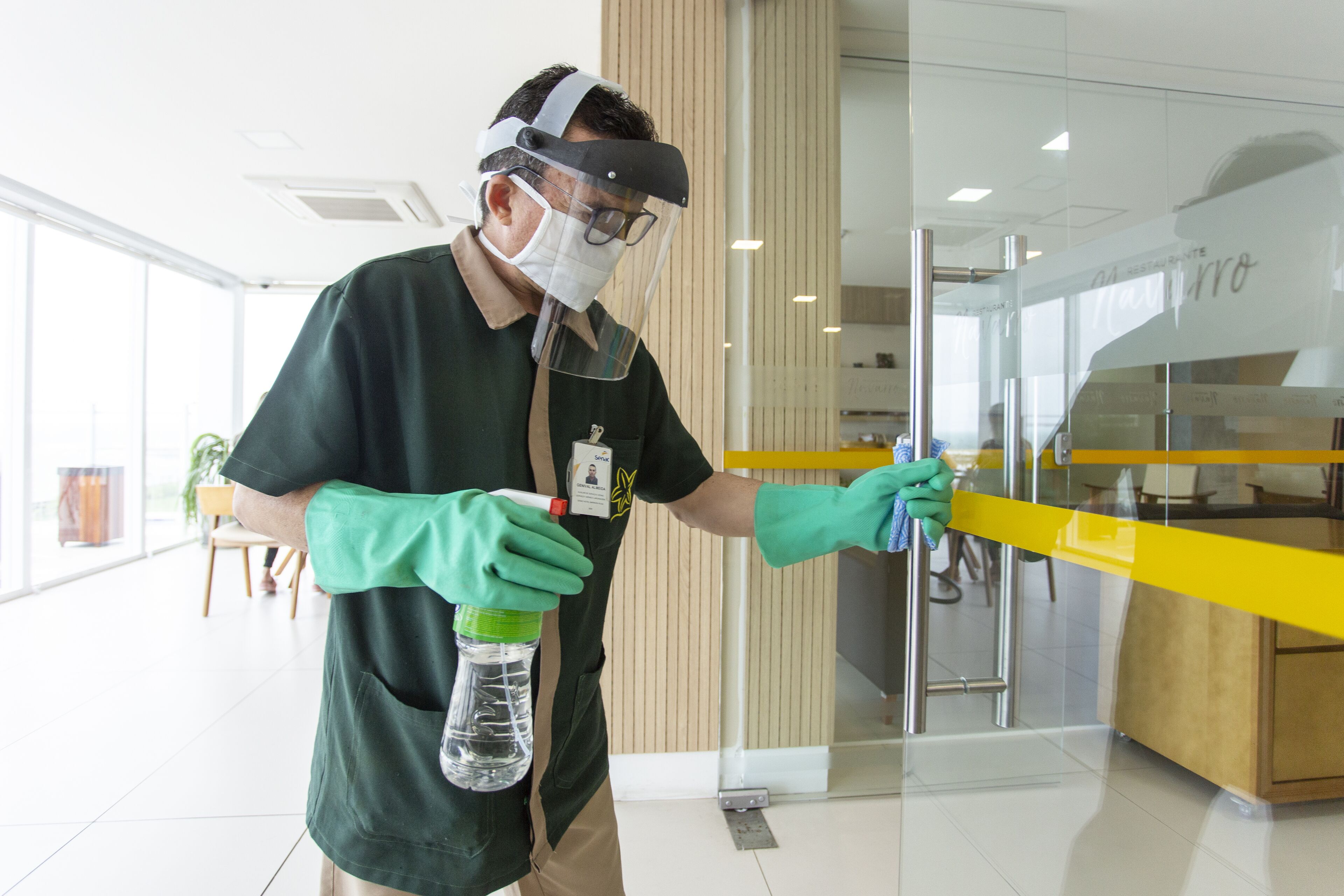 A person wearing a mask and gloves cleaning a glass partition in a modern interior space with wooden accents and a yellow counter.