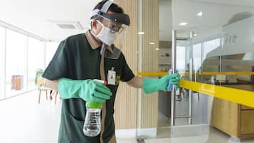 A person wearing a mask and gloves cleaning a glass partition in a modern interior space with wooden accents and a yellow counter.