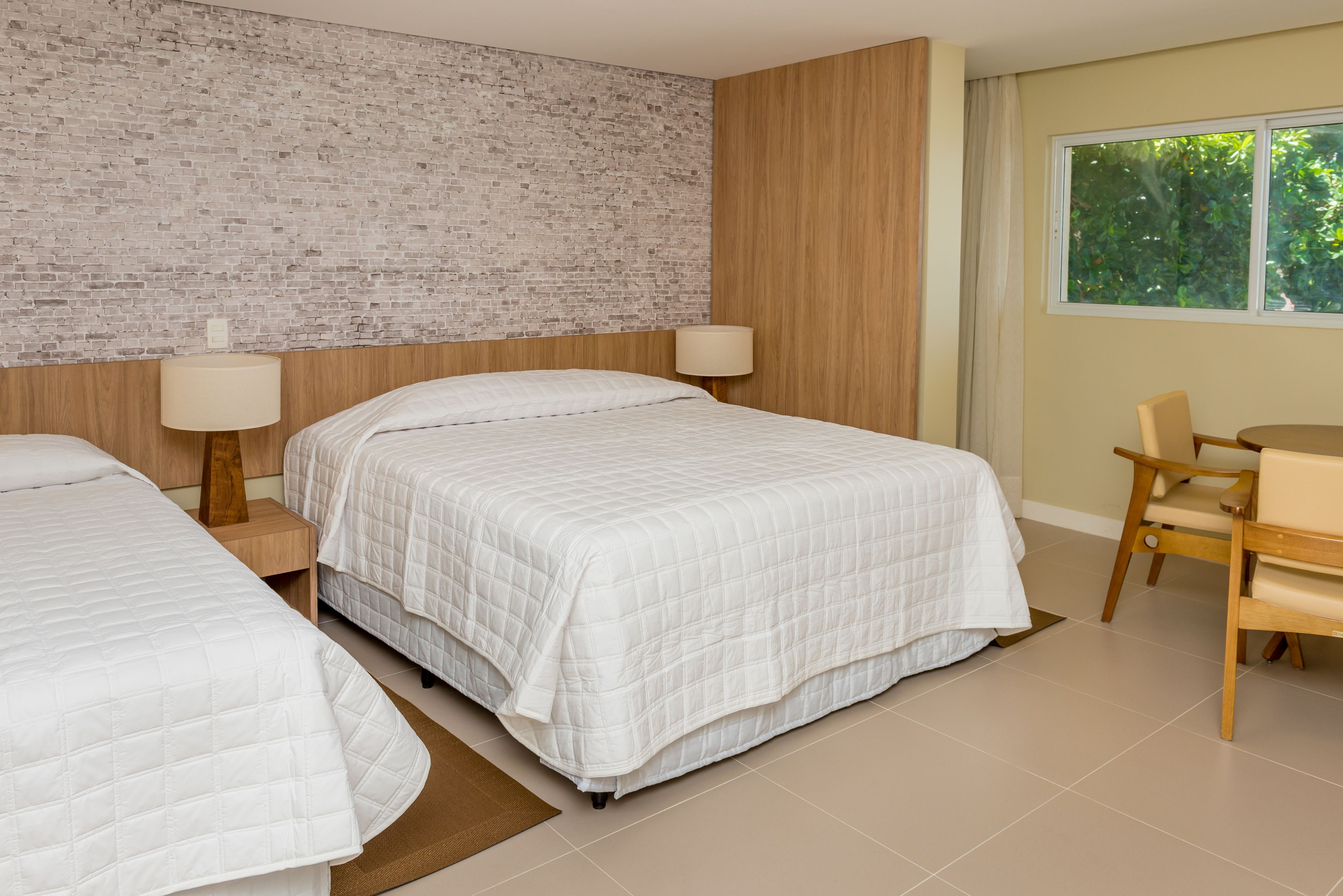 A hotel room with two beds, a brick wall, wooden paneling, a window with a view of greenery, and a desk with a chair.