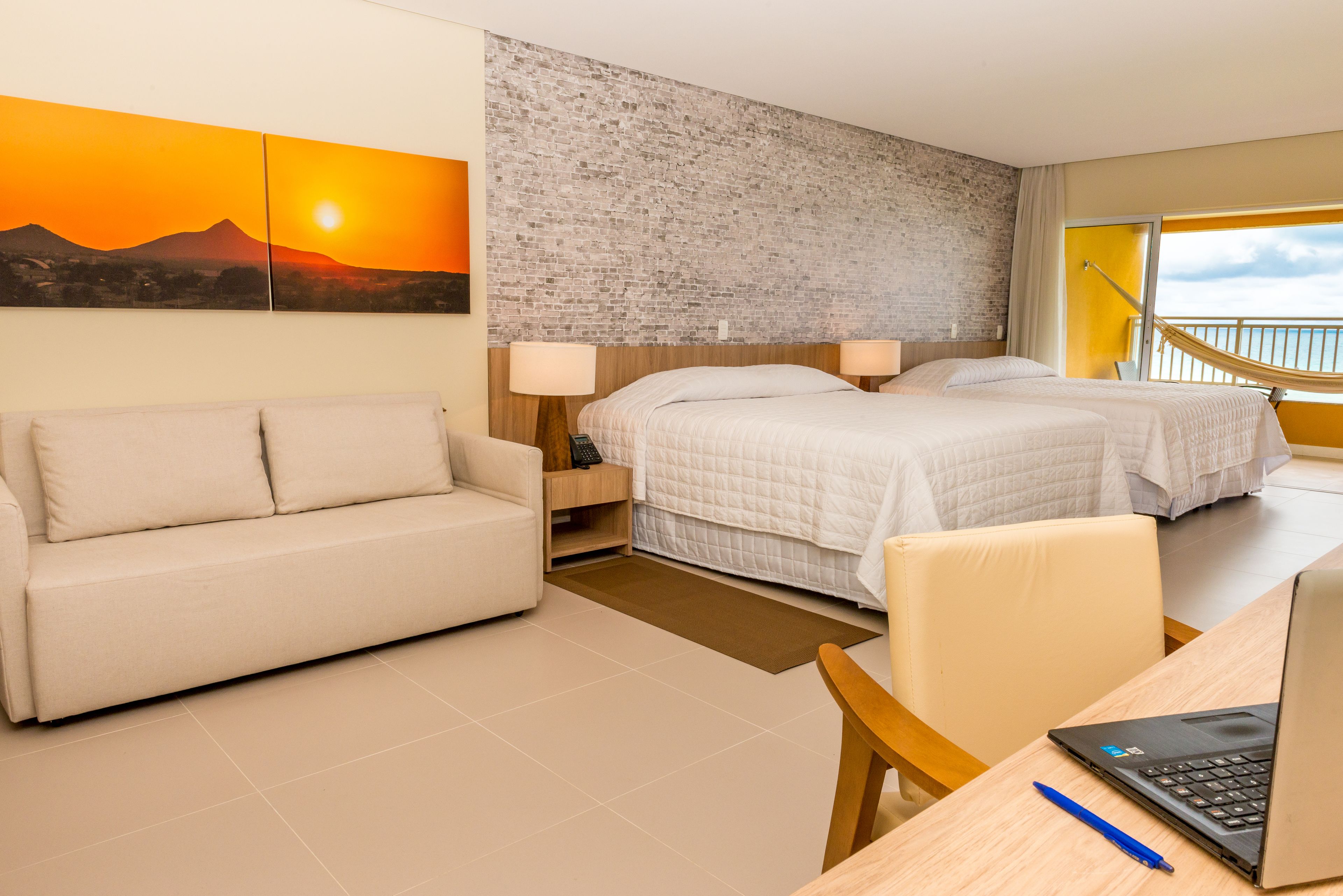 A hotel room with a bed, a sofa, a desk with a laptop, and a large window with a view of the sky.