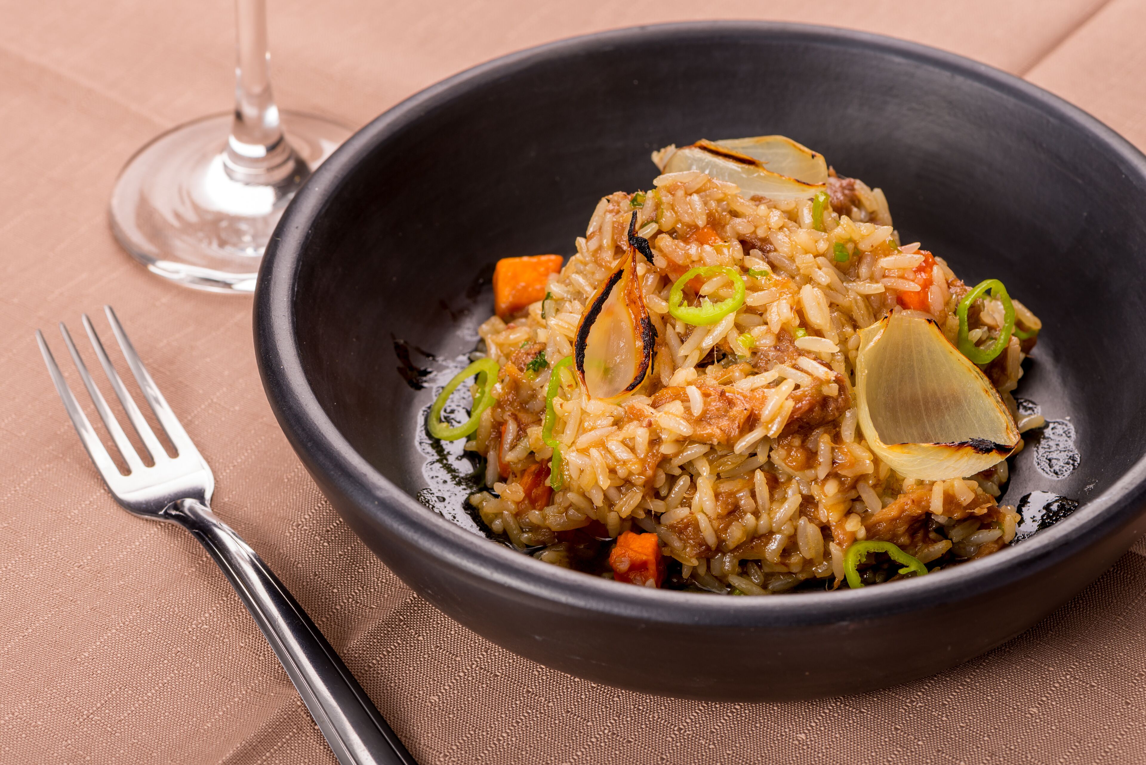 A bowl of fried rice with vegetables and meat, accompanied by a fork and a wine glass on a table.