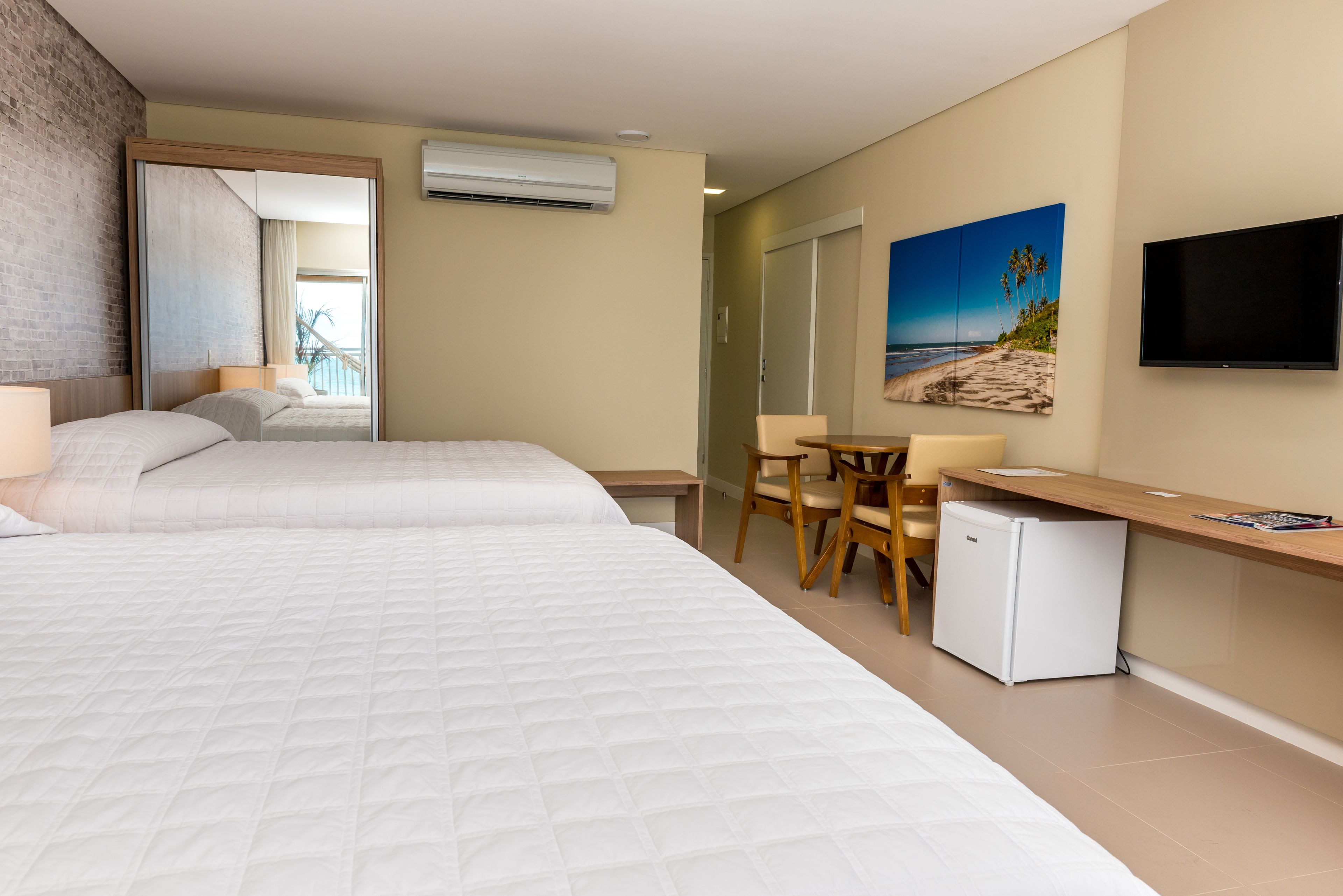 Habitación de hotel con dos camas, un escritorio, una silla, un refrigerador pequeño, un televisor de pantalla plana y un cuadro con motivos playeros en la pared.