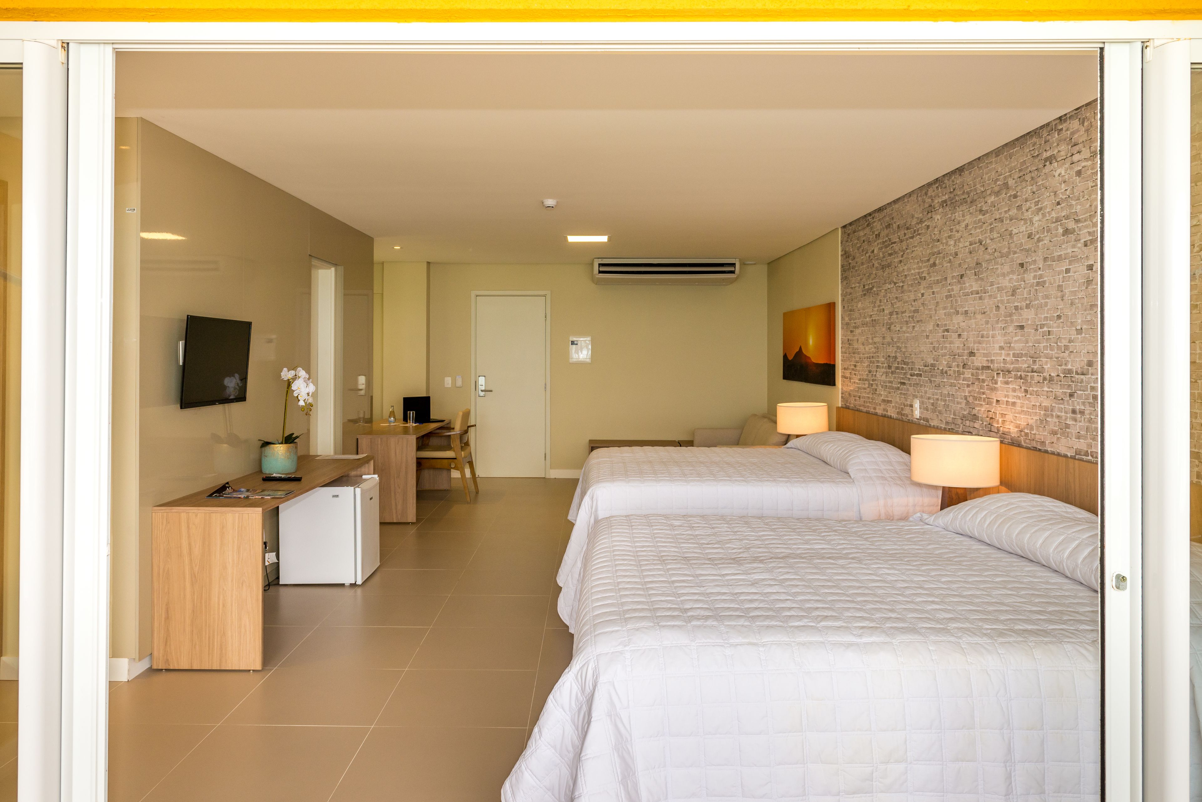 A hotel room with two beds, a desk, a chair, a TV, and a brick wall.