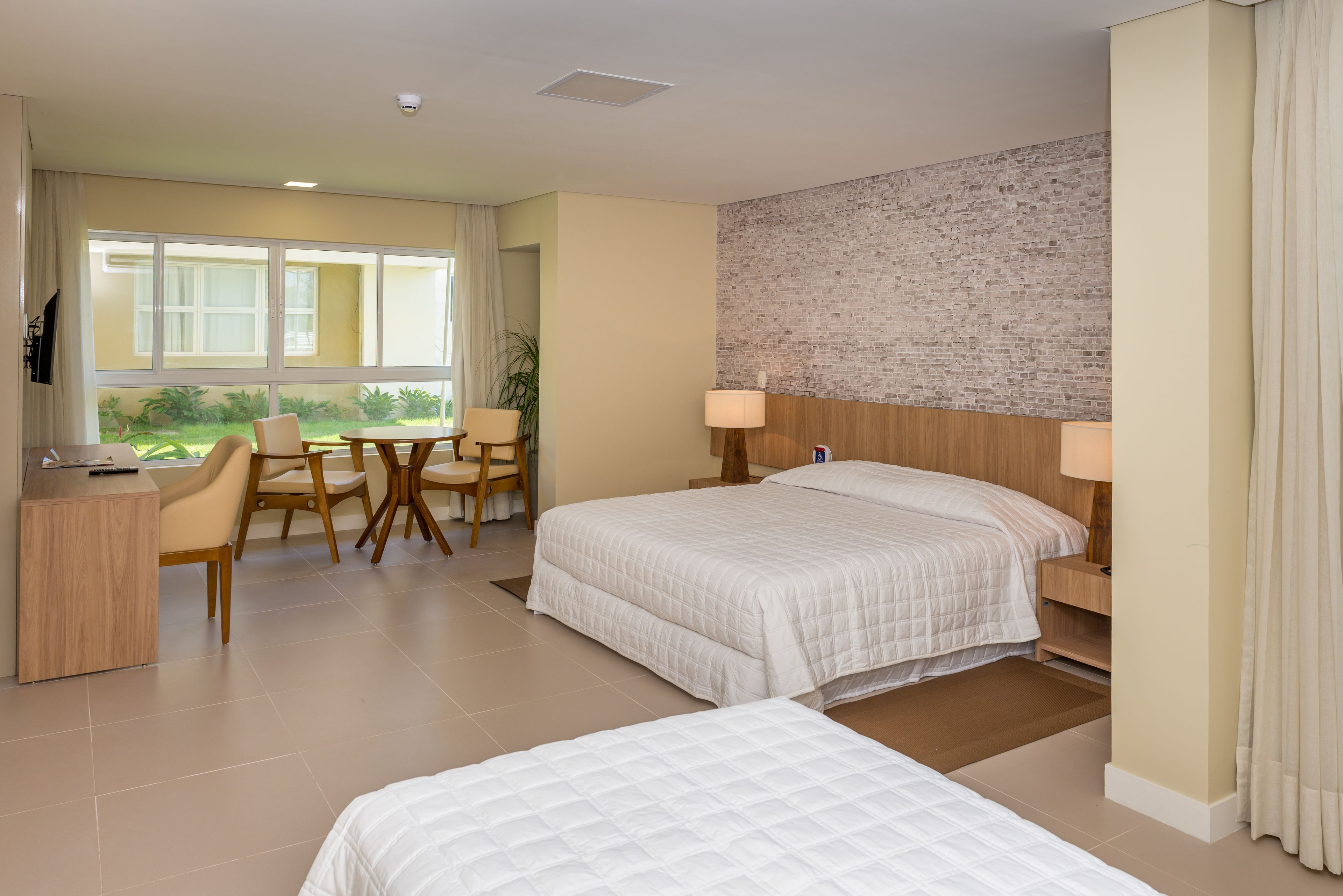 A hotel room with two beds, a desk, a chair, and a view of greenery outside.
