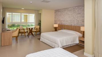 A hotel room with two beds, a desk, a chair, and a view of greenery outside.