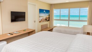 Ein Hotelzimmer mit zwei Betten, einem Fernseher, einem Strandbild und einem Fenster mit Meerblick.