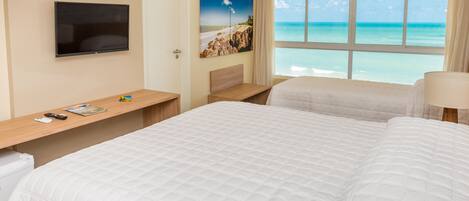 A hotel room with two beds, a TV, a painting of a beach, and a window with a view of the ocean.