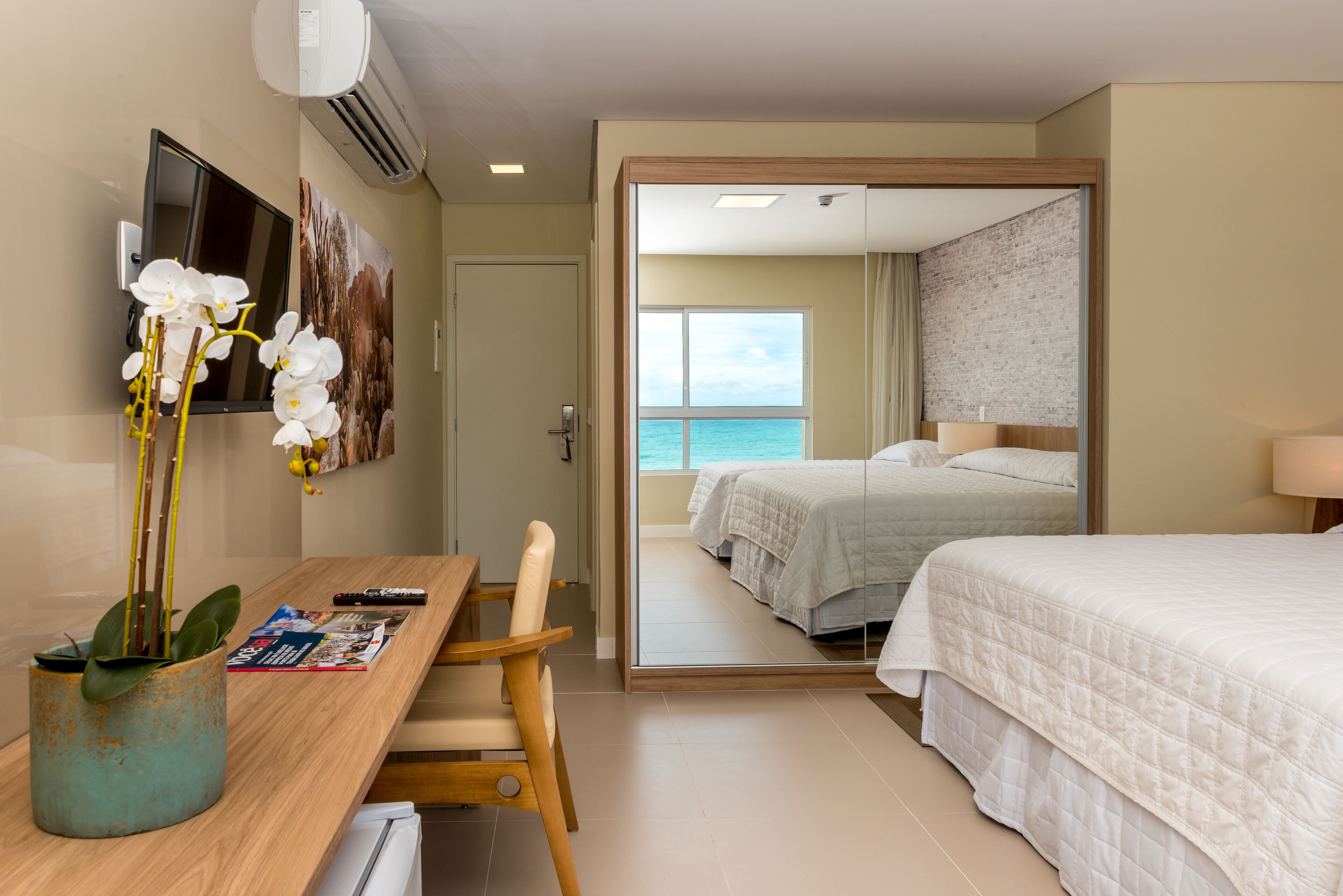 Habitación de hotel con un escritorio de madera, una orquídea en maceta, una cama con ropa de cama blanca, un ventanal con vistas al mar y un espejo que refleja la habitación.