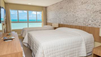 A hotel room with two beds, a wooden desk, a TV, and a large window with a view of the ocean.