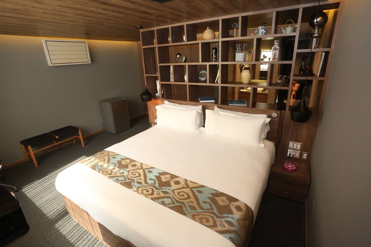 Ein ordentlich eingerichtetes Hotelzimmer mit einem großen Bett, einem Nachttisch, einer eingebauten Ablage mit verschiedenen Gegenständen und einer Bank in der Ecke.
