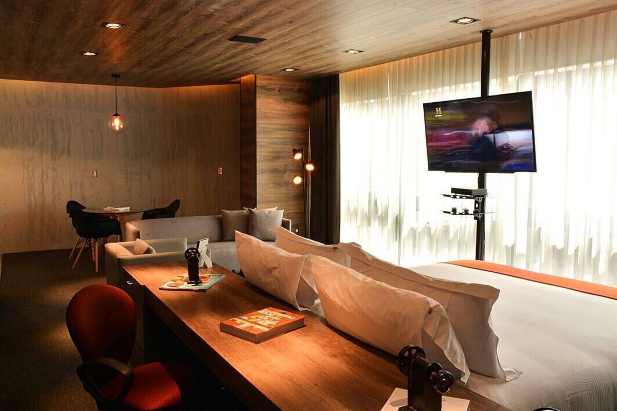 Ein modernes Wohnzimmer mit Holzbalkendecke, einem großen Flachbildfernseher, einer Couch, einem Esstisch mit Stühlen und einem großen Fenster mit transparenten Vorhängen.
