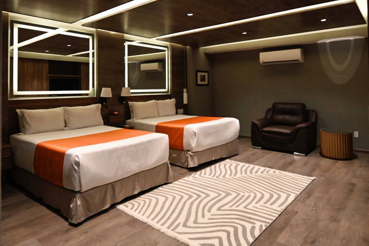 Ein modernes Hotelzimmer mit zwei Betten, einem Ledersessel, einem großen Spiegel und einem gemusterten Teppich.