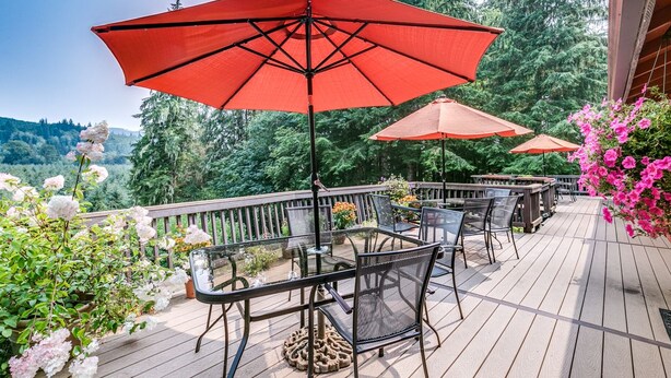 Una terraza de madera con mobiliario exterior y sombrillas rojas, rodeada de abundante follaje y flores.