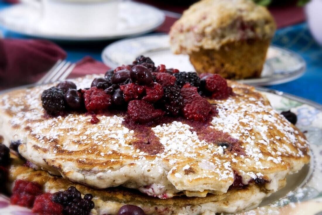Una pila de tortitas cubiertas de frutos rojos y azúcar glas, con un muffin al fondo.