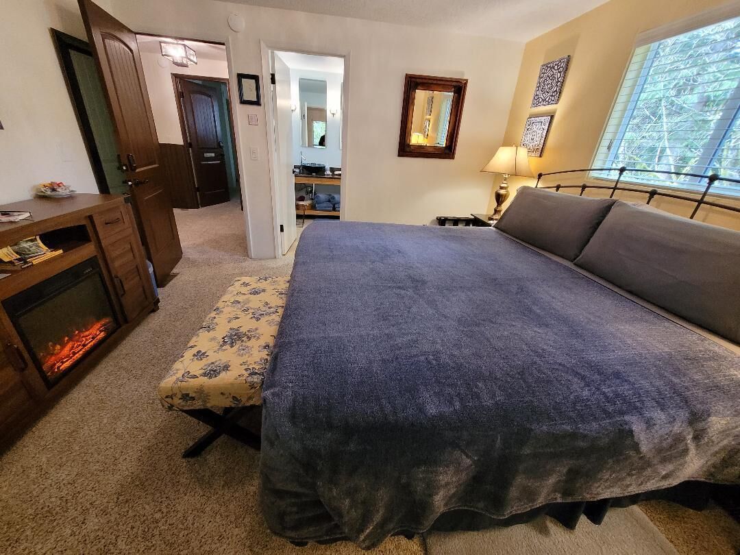 A bedroom with a large bed, a bedside table, a lamp, a fireplace, and a door leading to another room.