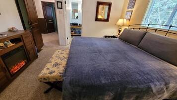 A bedroom with a large bed, a bedside table, a lamp, a fireplace, and a door leading to another room.